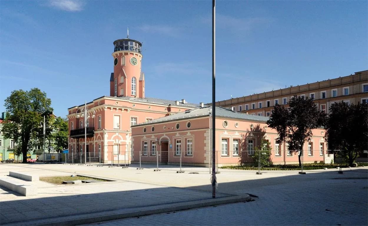 A large, light pink building with a prominent clock tower stands in the center of a spacious paved plaza under a clear blue sky, with a larger, tan building visible in the background.